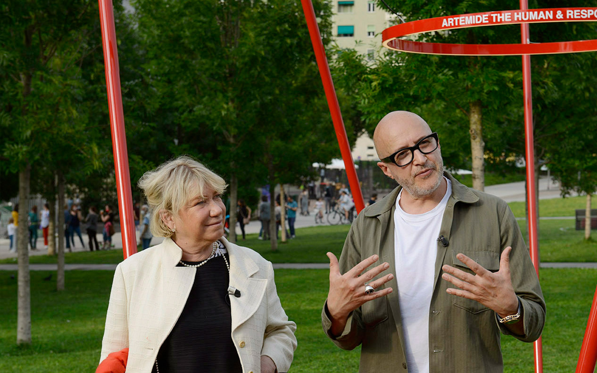 Two people in a park discussing near a red Artemide installation with the text 'Artemide The Human & Respo...'