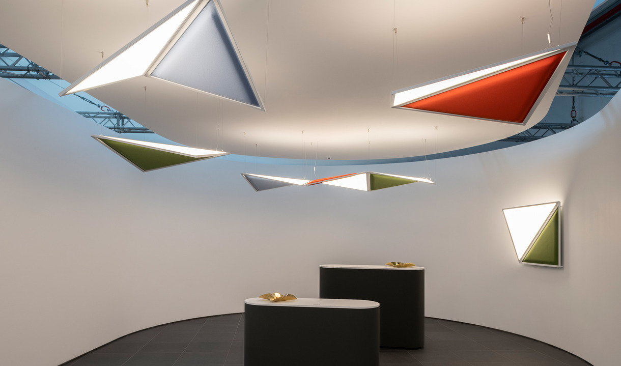 Triangular pendant and wall lights in white, red, green, and blue hang in a curved white room above two dark reception desks.
