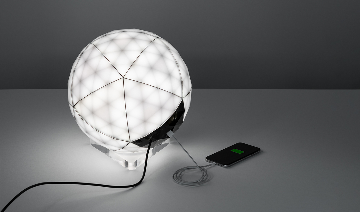 Artemide spherical LED table lamp with geometric faceted diffuser, resting on transparent base, with USB charging port and connected smartphone