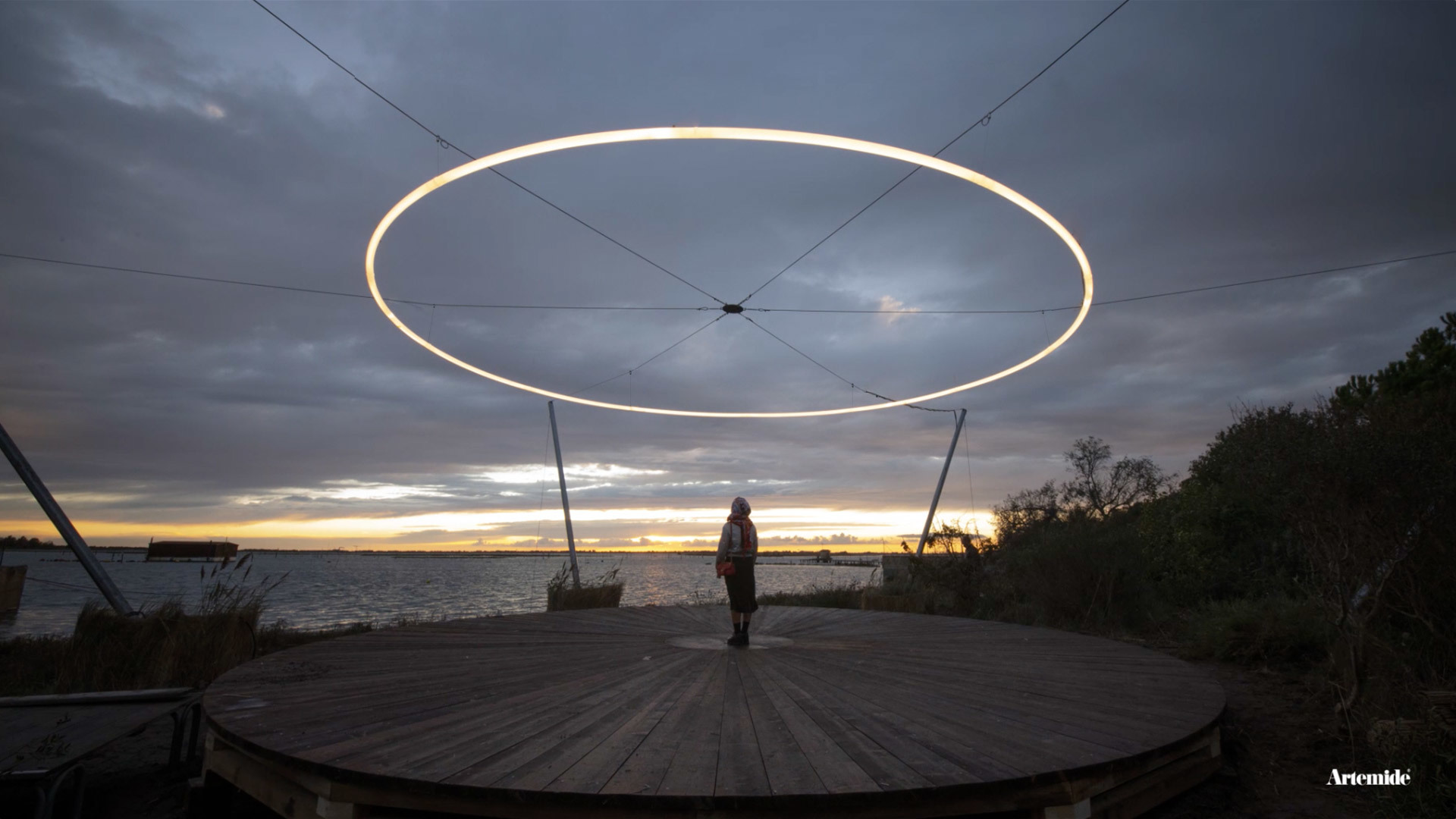 Person standing on a wooden deck beneath a large illuminated circular Artemide ring light suspended outdoors at dusk over a waterfront.