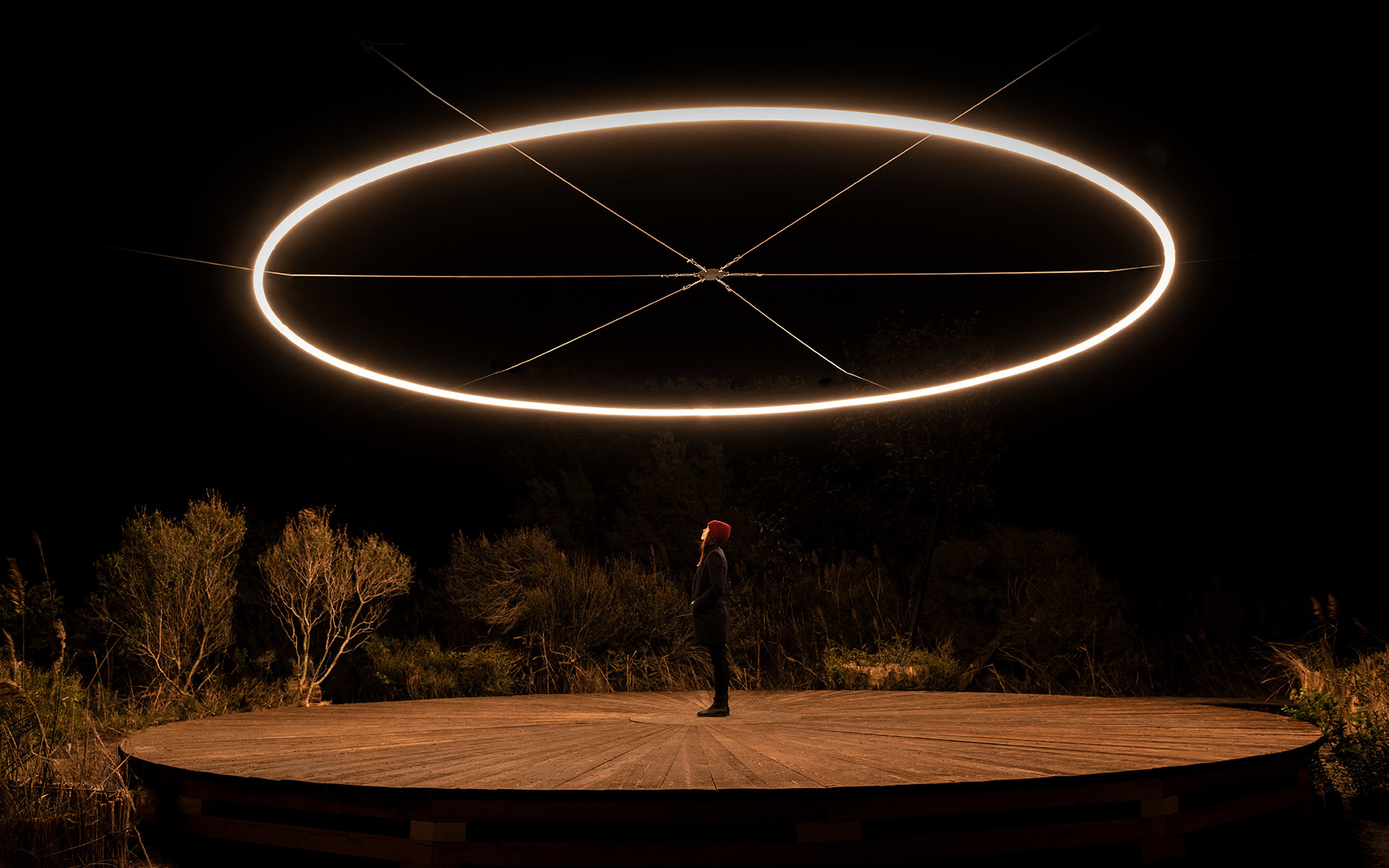 Large glowing elliptical ring light suspended outdoors at night over a circular wooden platform with a person standing beneath it