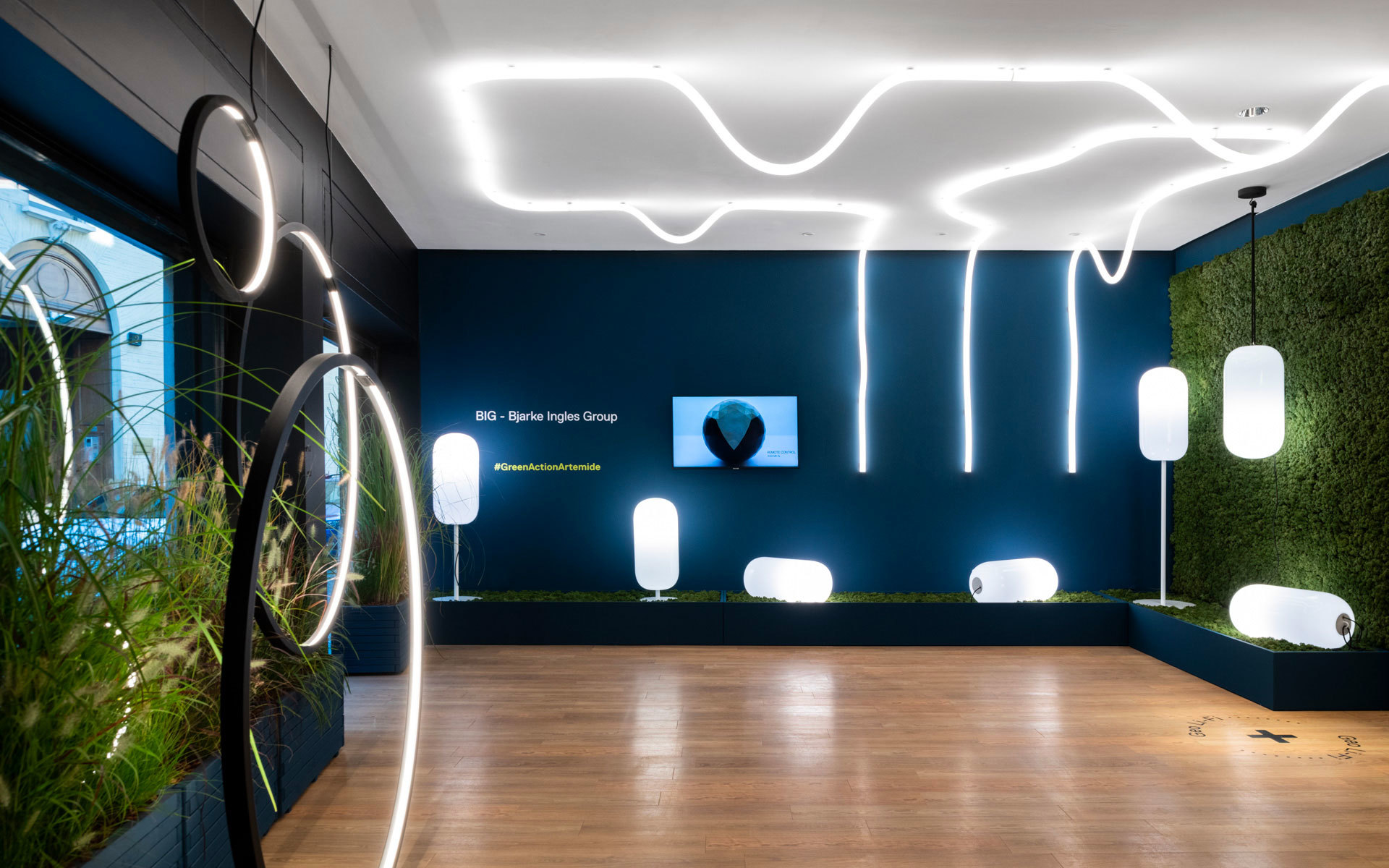 Artemide showroom with BIG collection: organic LED ceiling lines, circular floor lamps, and white oval pendant lights against teal wall with greenery.