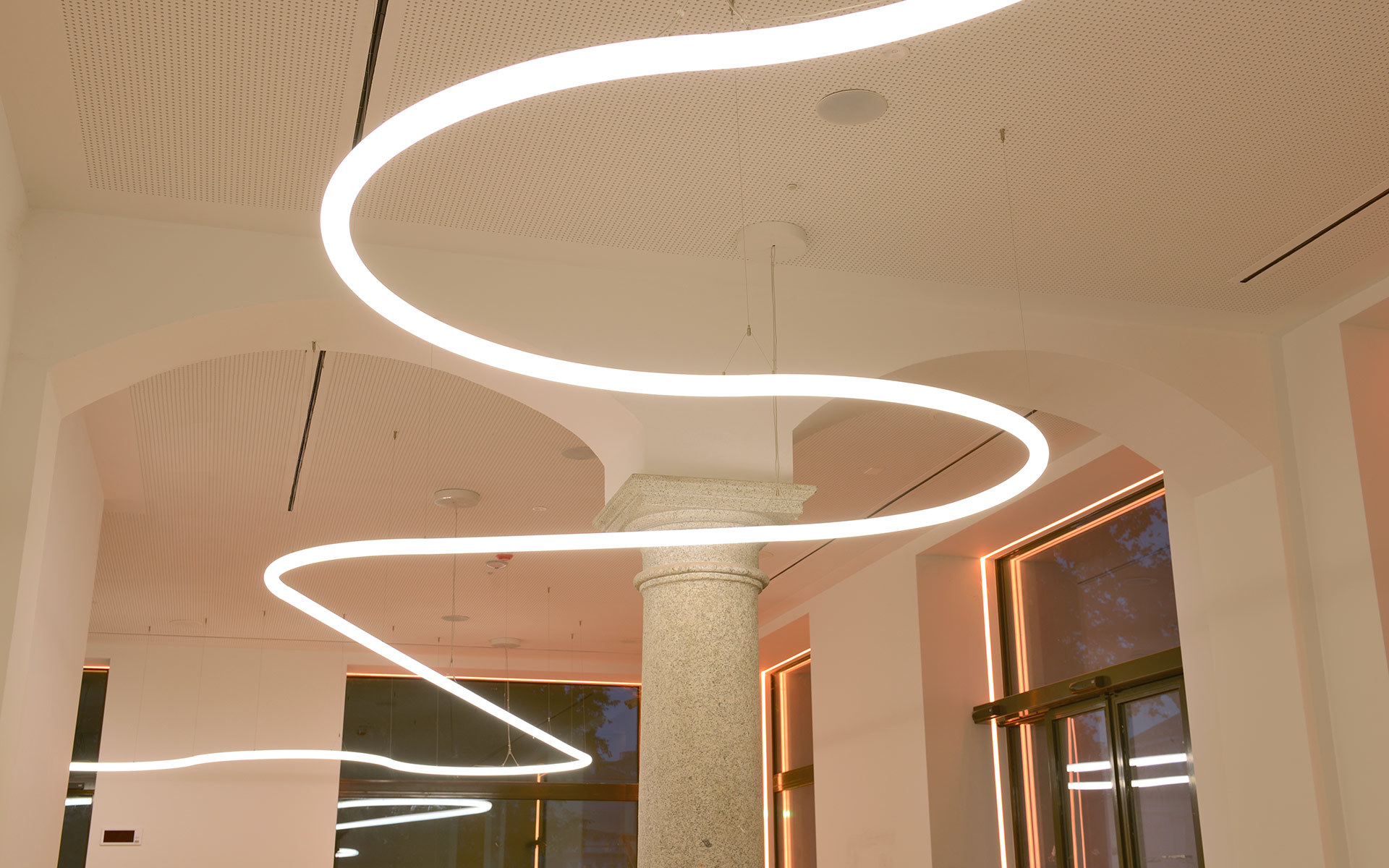 Curved freeform LED pendant light installations suspended from a white ceiling above a classical column in an elegant interior space.