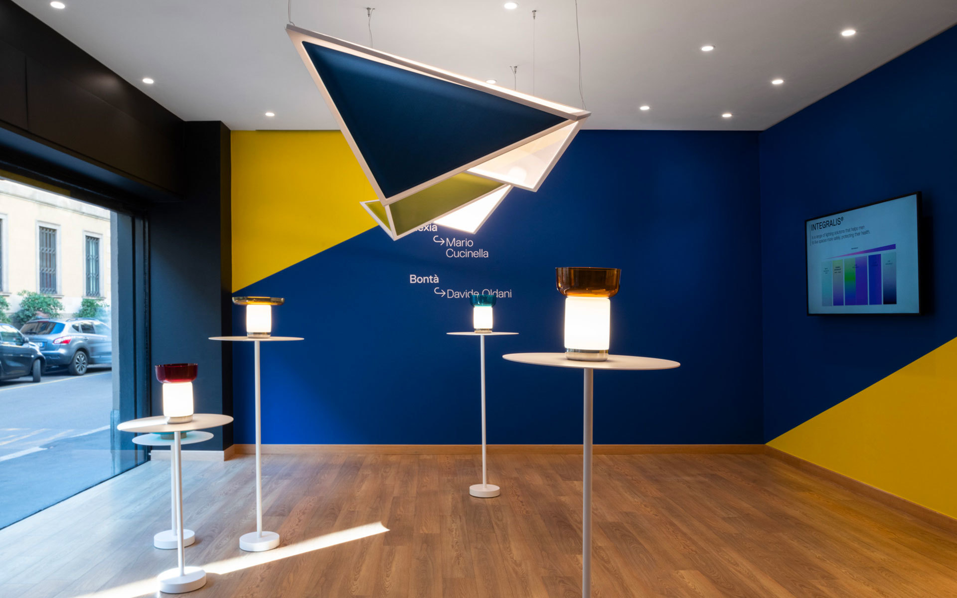 Artemide showroom with geometric pendant light and Bontà table lamps by Davide Groppi on stands, against bold blue and yellow wall
