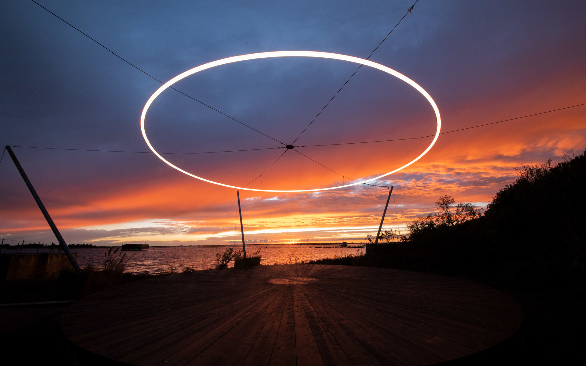 Large luminous white ring suspended outdoors against a dramatic red and orange sunset sky over water, supported by cables