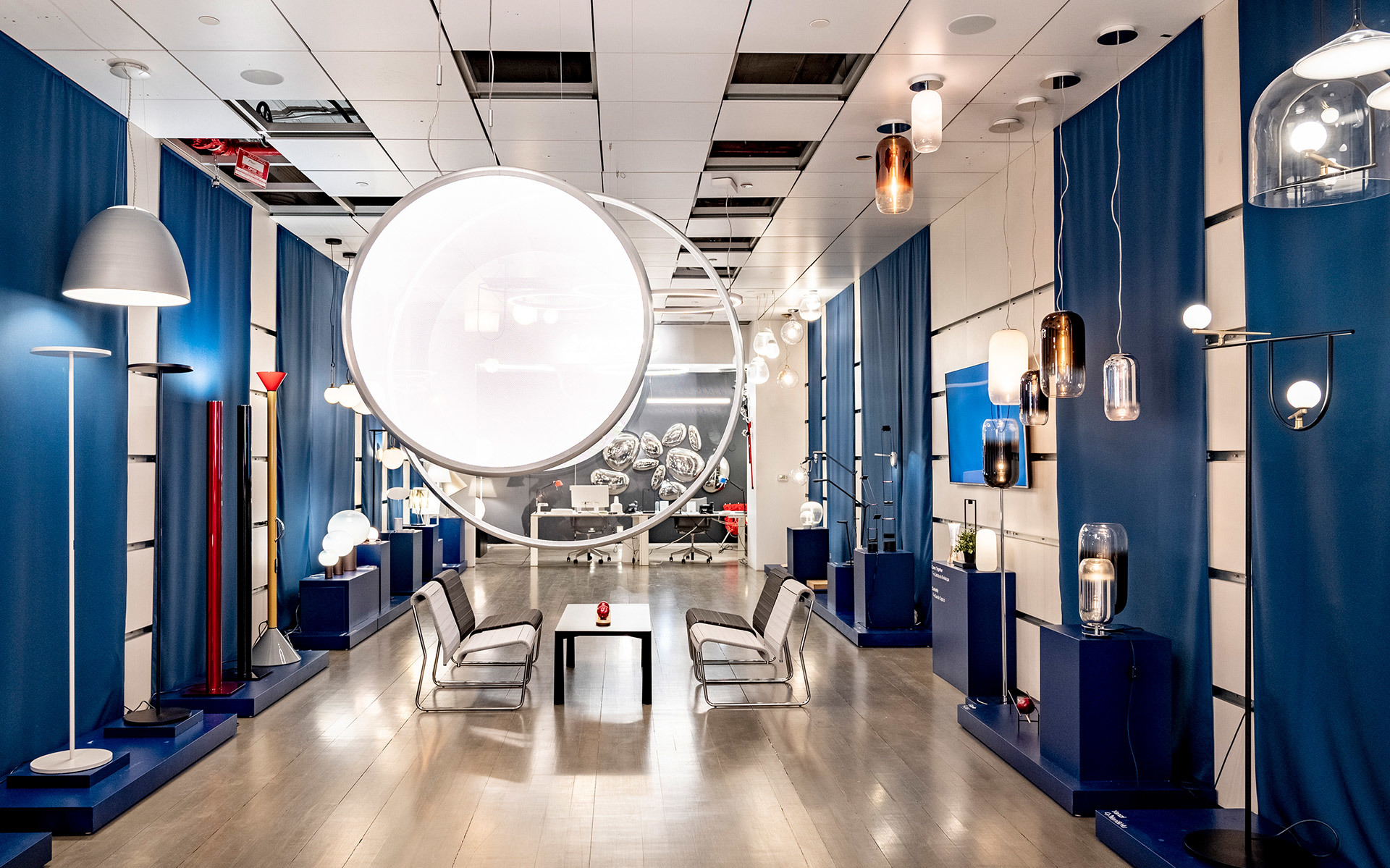 Artemide showroom with pendant, floor, and wall lamps displayed along blue-curtained walls, centered by a large circular light installation.