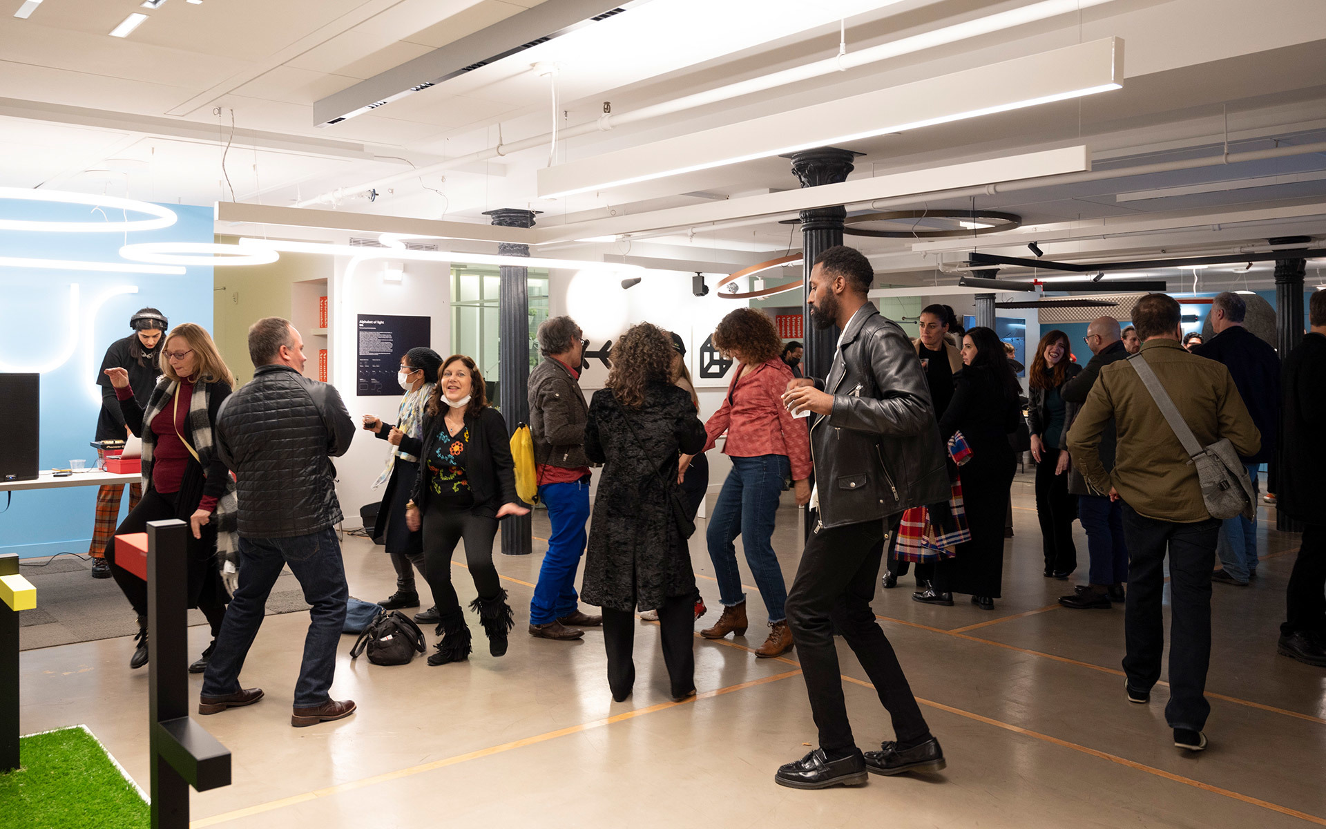 Crowded indoor event space with people mingling, illuminated by recessed and linear ceiling lights in a modern open-plan area.