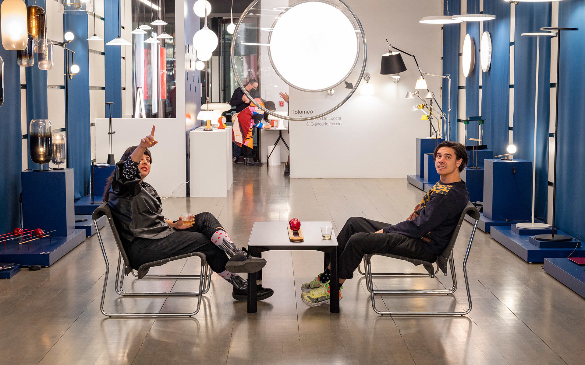 Two people seated facing each other in an Artemide showroom surrounded by pendant, floor, and wall lamps on blue displays.