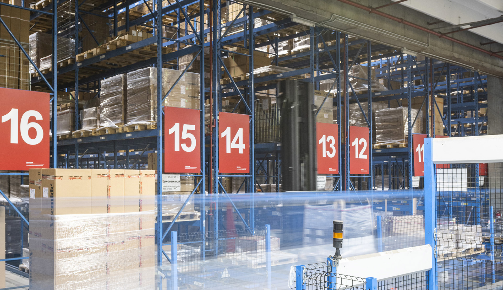 Large warehouse interior with numbered blue metal shelving racks (11-16) storing palletized goods on multiple levels.