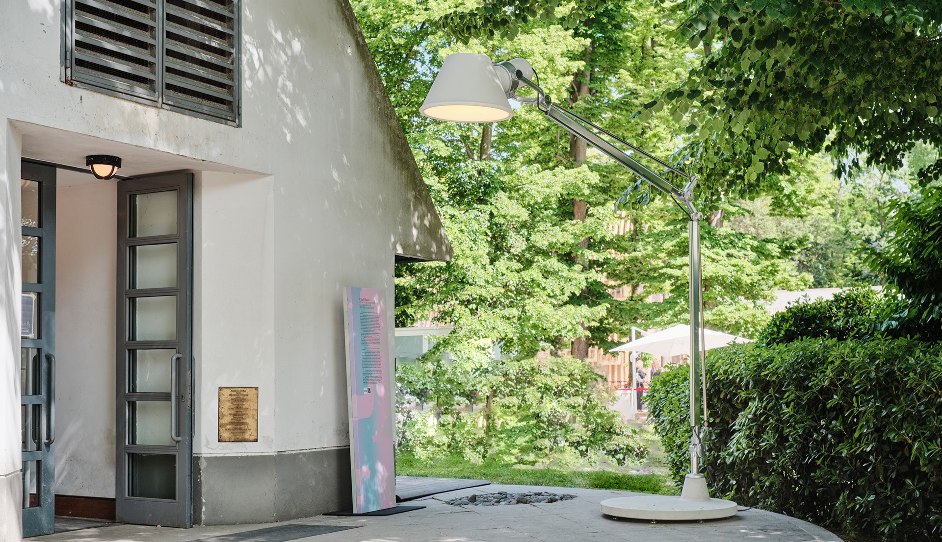 Artemide Tolomeo Mega Terra floor lamp in aluminum, placed outdoors near a building entrance surrounded by lush green trees.