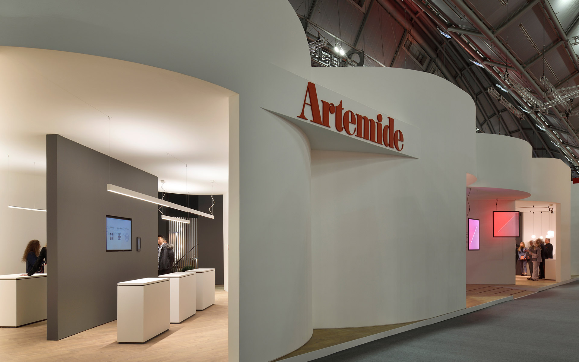 Artemide exhibition stand with white curved walls, logo, and a dark interior showroom displaying pendant and linear lighting fixtures.