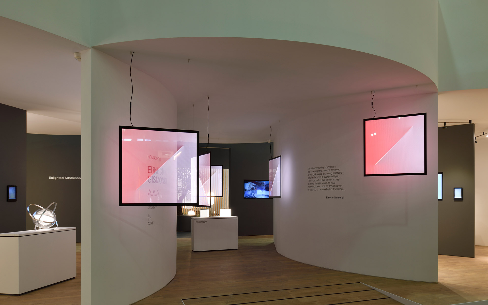 Modern exhibition space with curved white walls, suspended digital screens displaying pink visuals, and a sculptural white lamp on a pedestal.