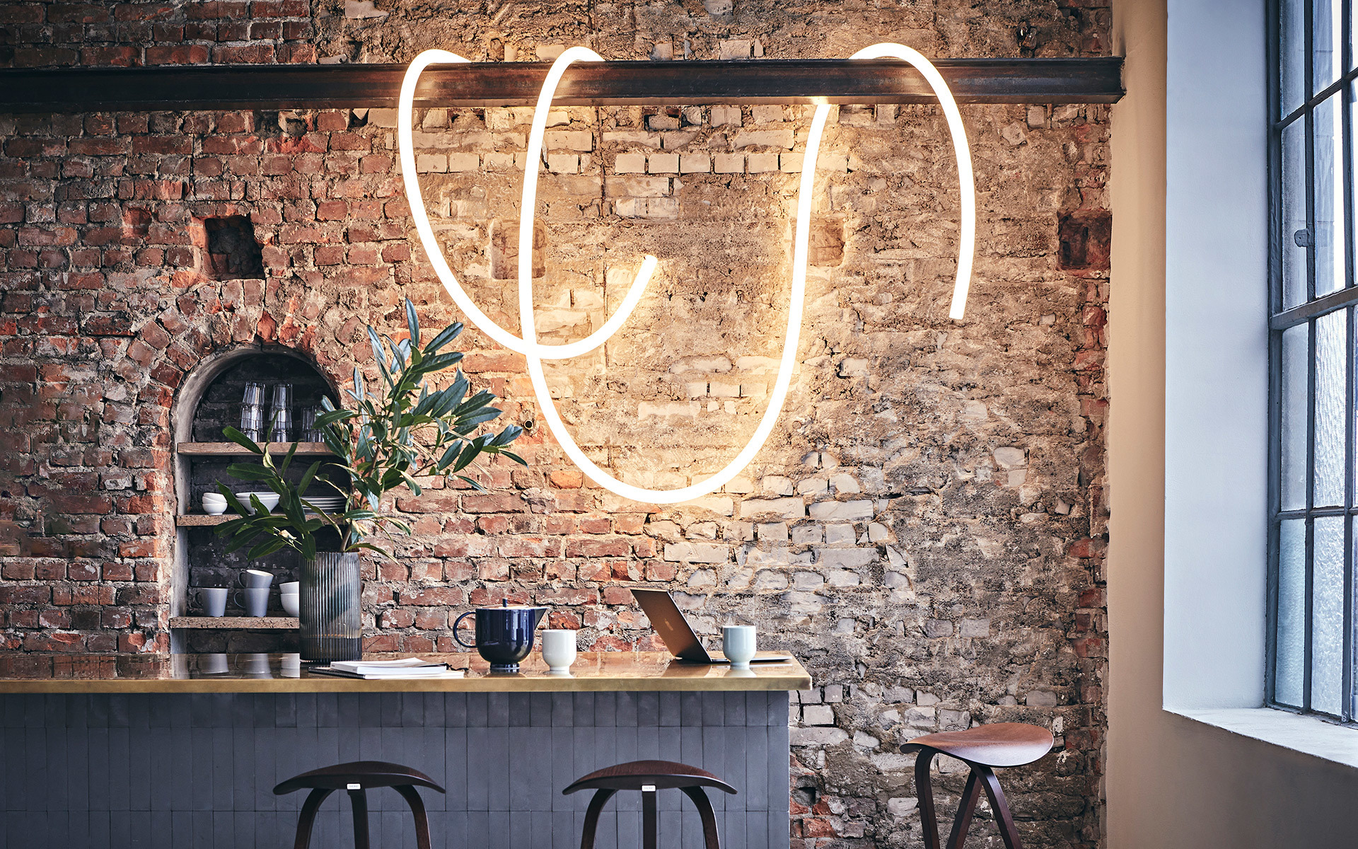 Glowing neon light sculpture in curved shapes mounted on exposed brick wall above a wooden bar counter with stools.