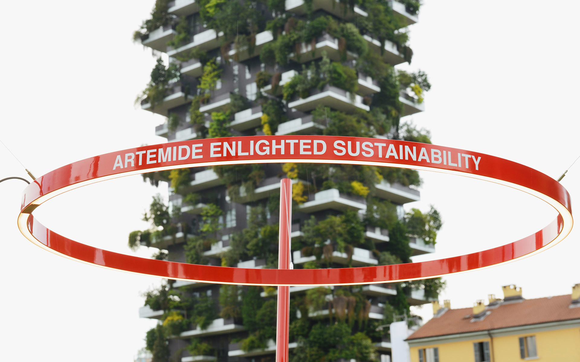 Red circular ring installation with 'Artemide Enlightened Sustainability' text, displayed in front of Milan's Bosco Verticale tower.
