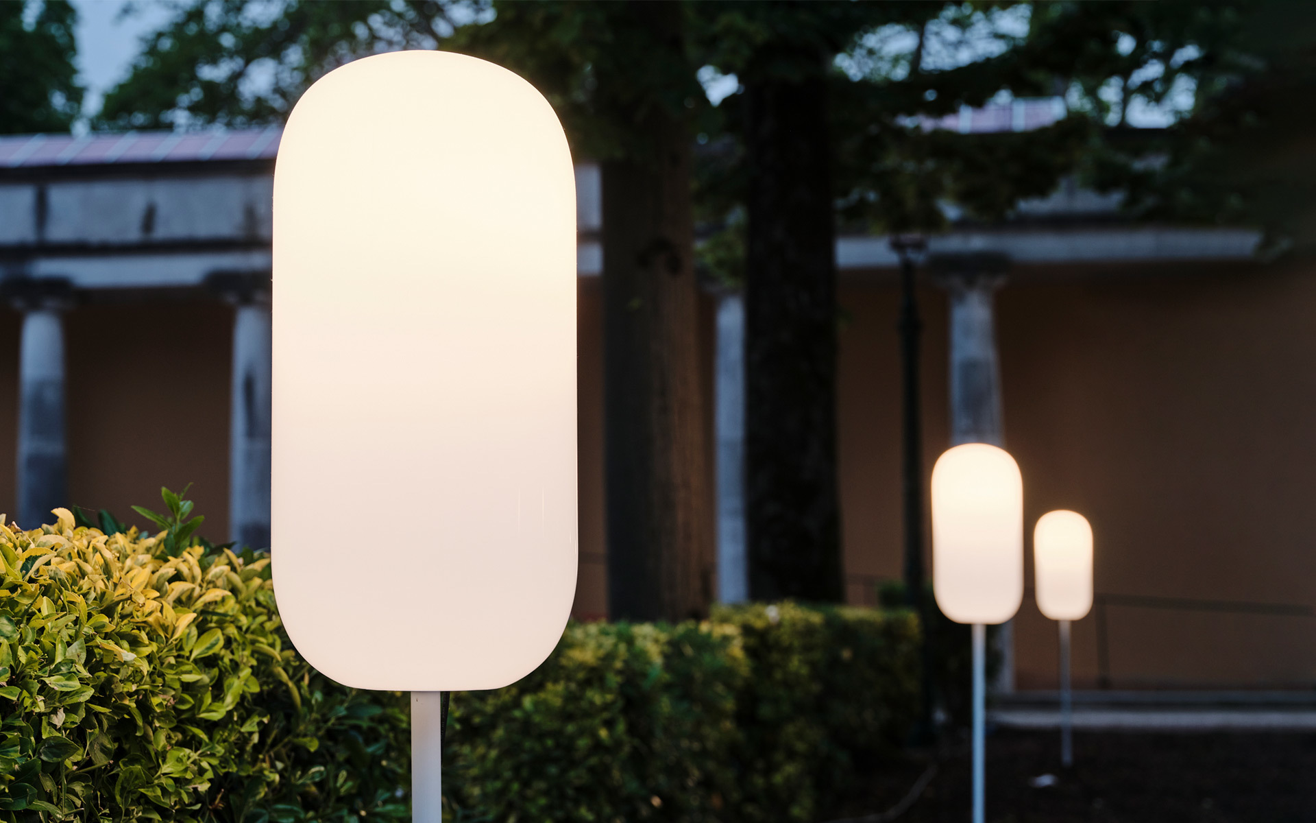 Three white opal pill-shaped outdoor floor lamps on slender poles, glowing softly at dusk in a garden setting
