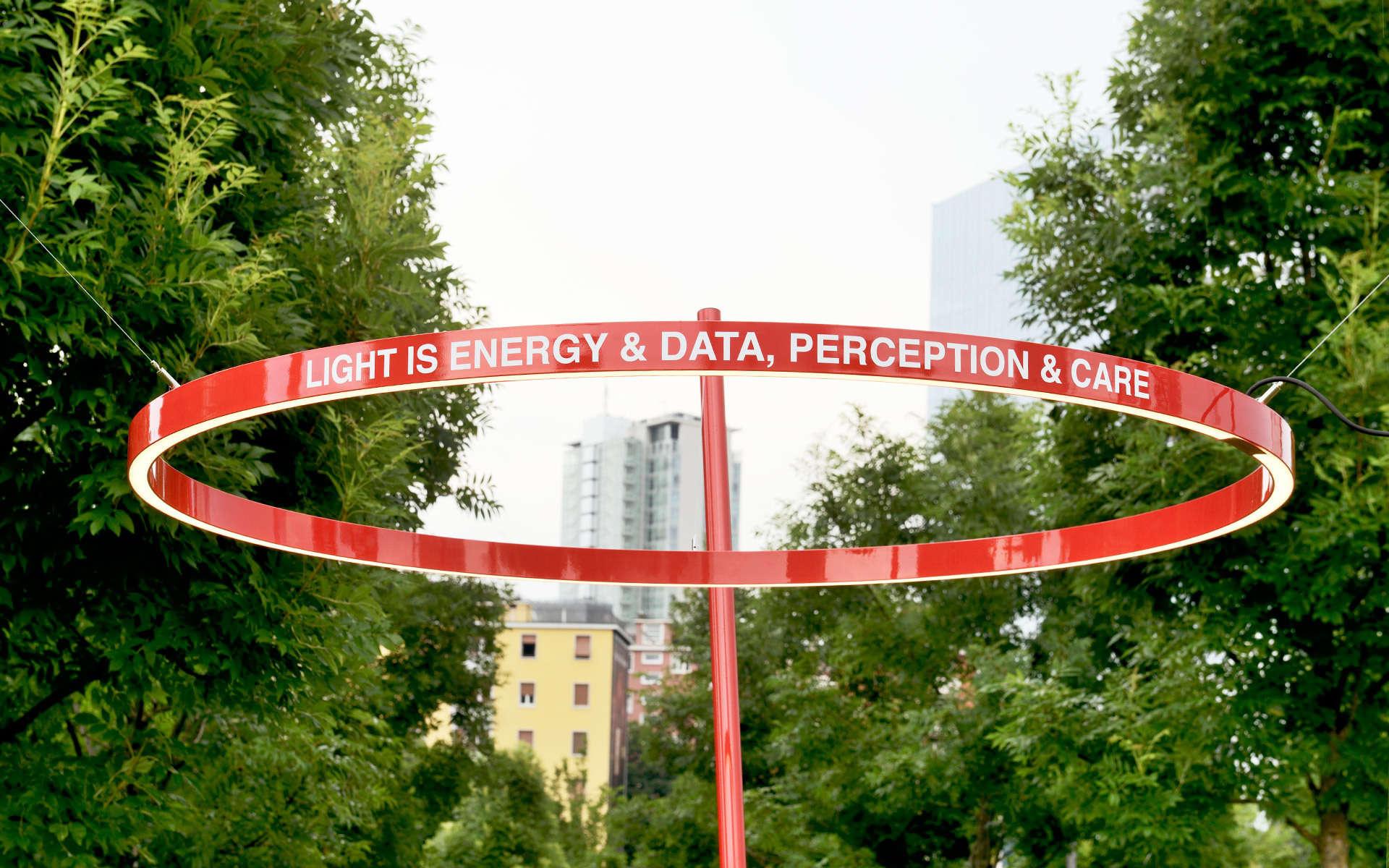 Red circular installation on a pole with the phrase 'Light is Energy & Data, Perception & Care' against a green tree background.