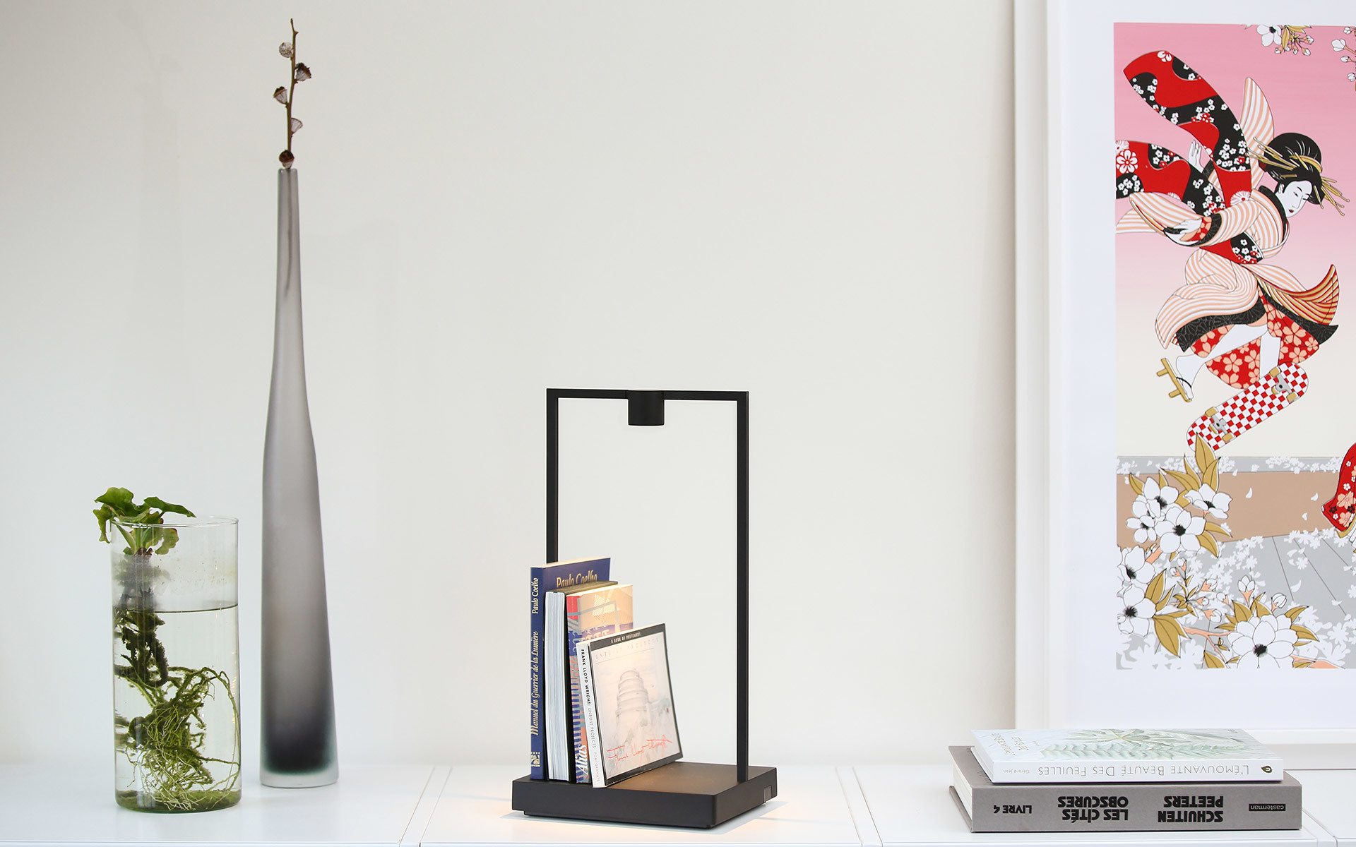 Black rectangular frame table lamp with wooden base holding small books, displayed on a white shelf with decorative vases and artwork.
