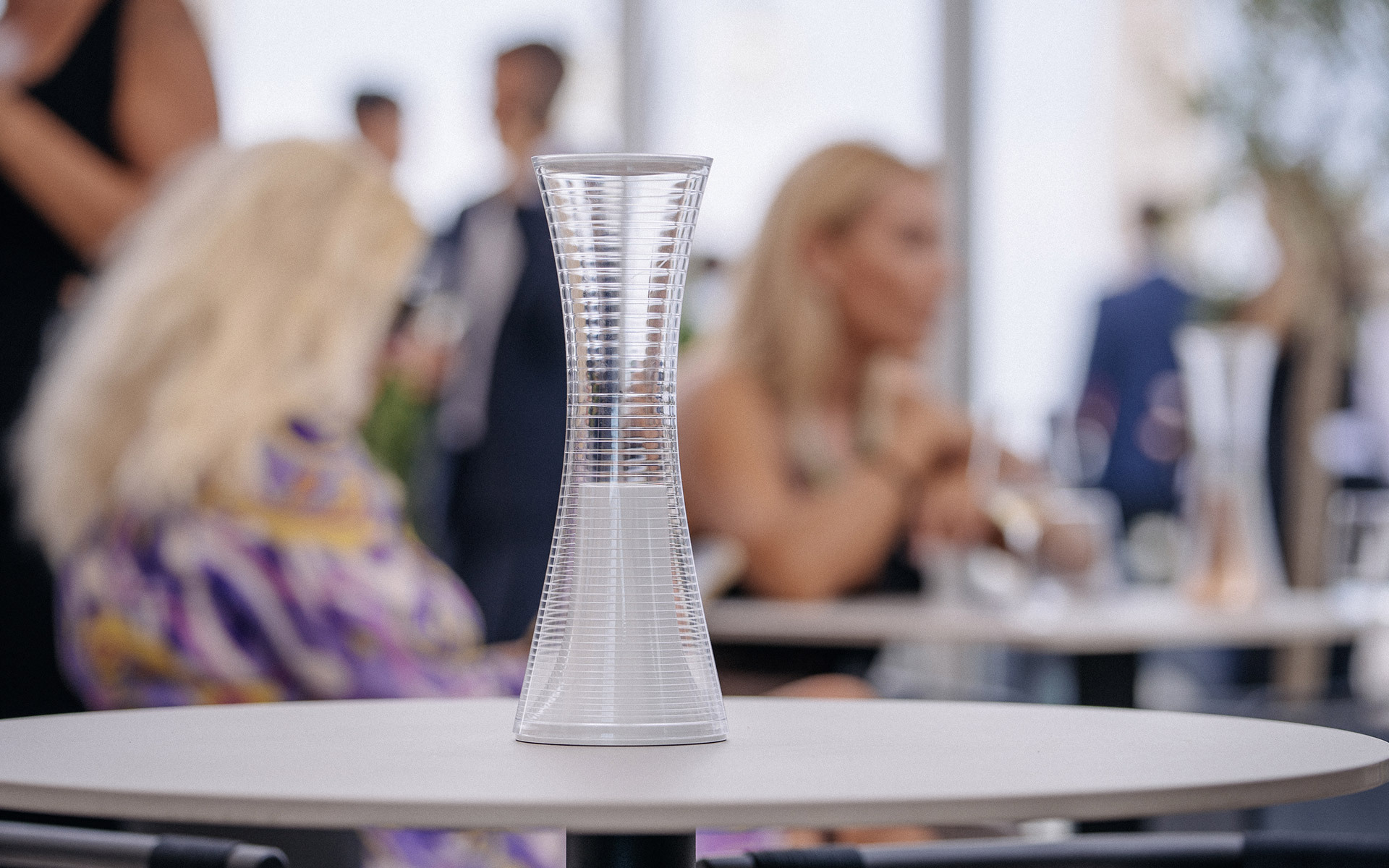 qelvornixuta.onlinee Together portable table lamp, hourglass-shaped transparent diffuser, displayed on a white table at an elegant social event
