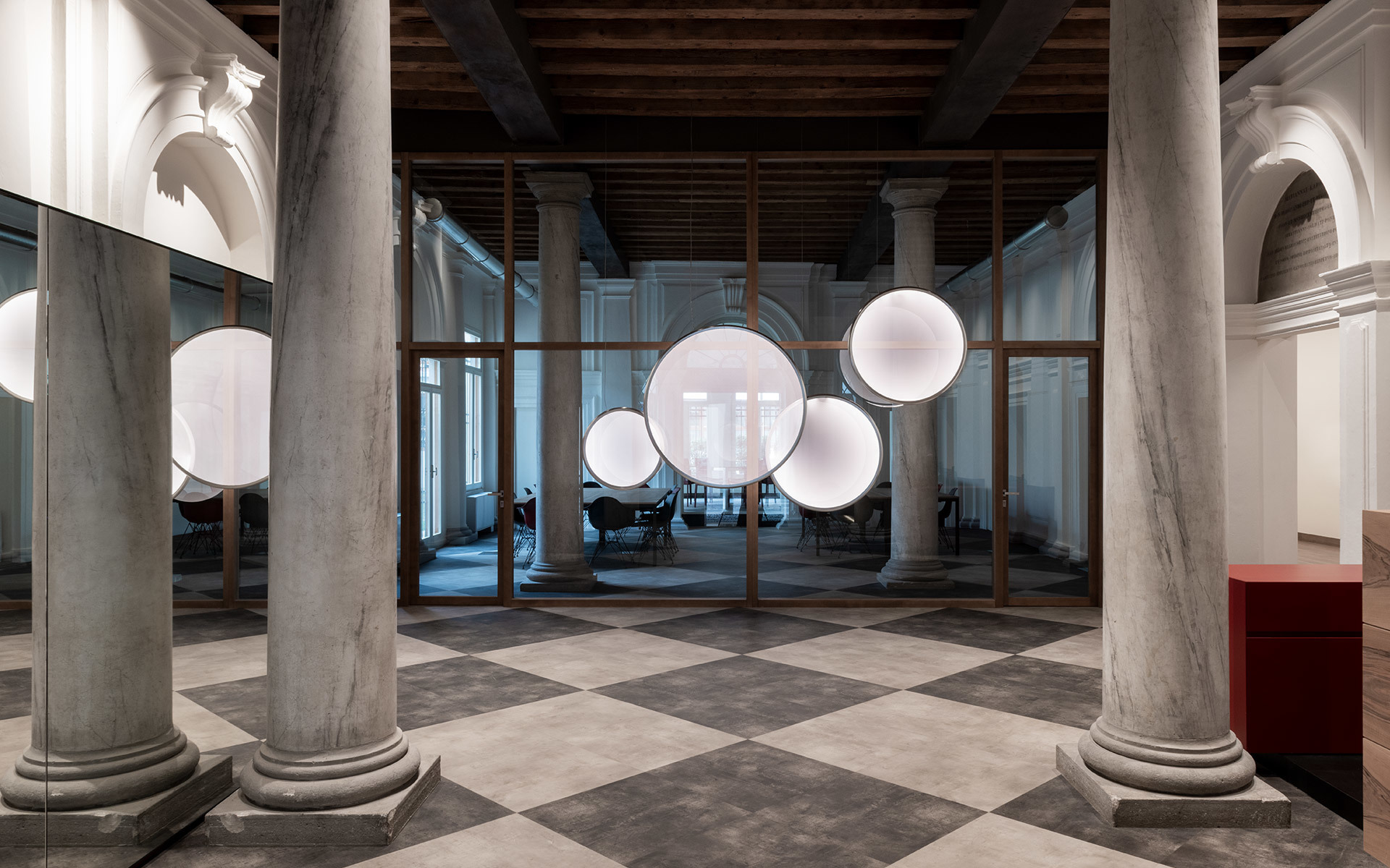 Cluster of glowing spherical pendant lamps suspended in a historic hall with marble columns and checkered floor.