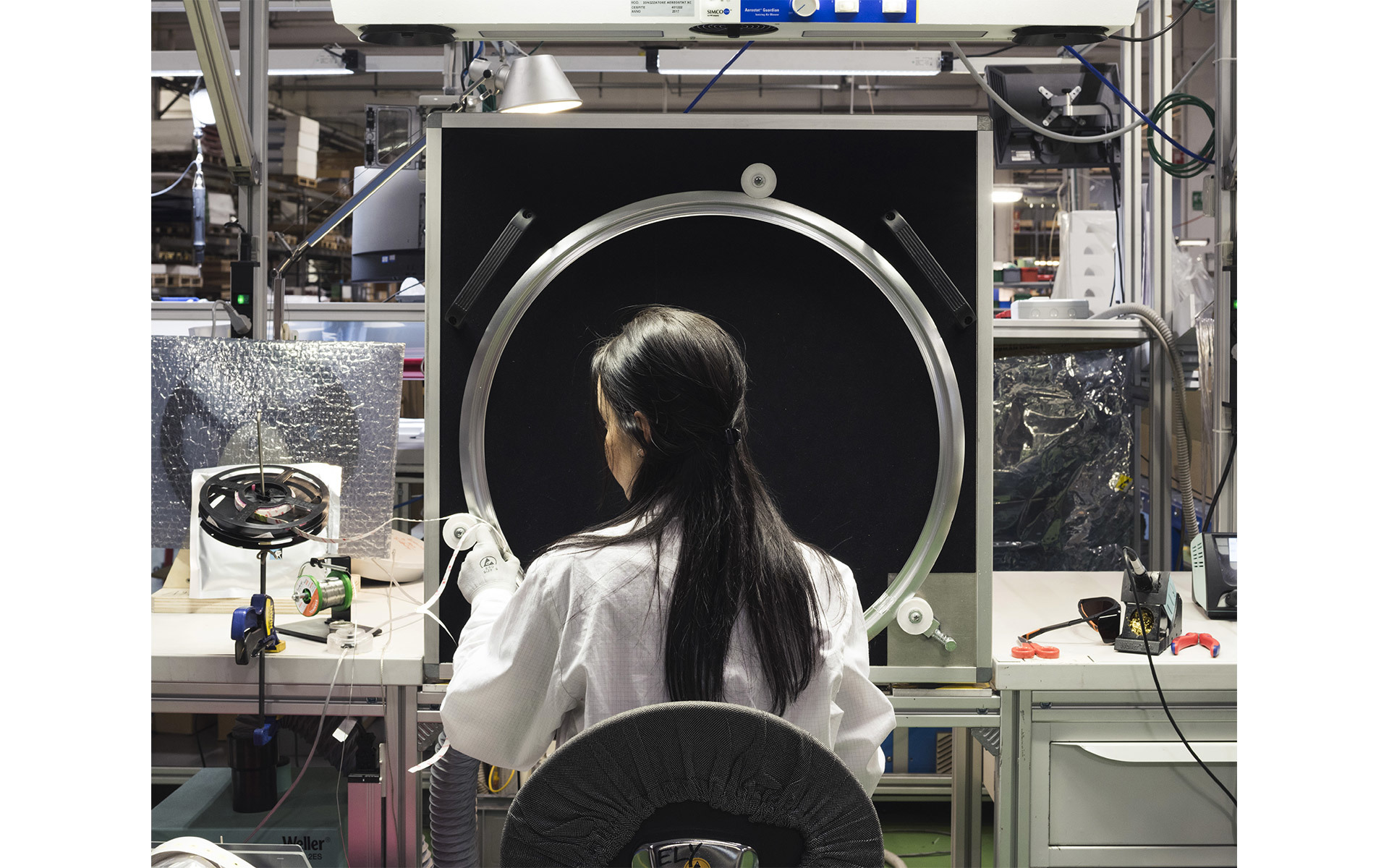 Technician in white coat assembling a large circular Artemide lamp component on a production line in a manufacturing facility