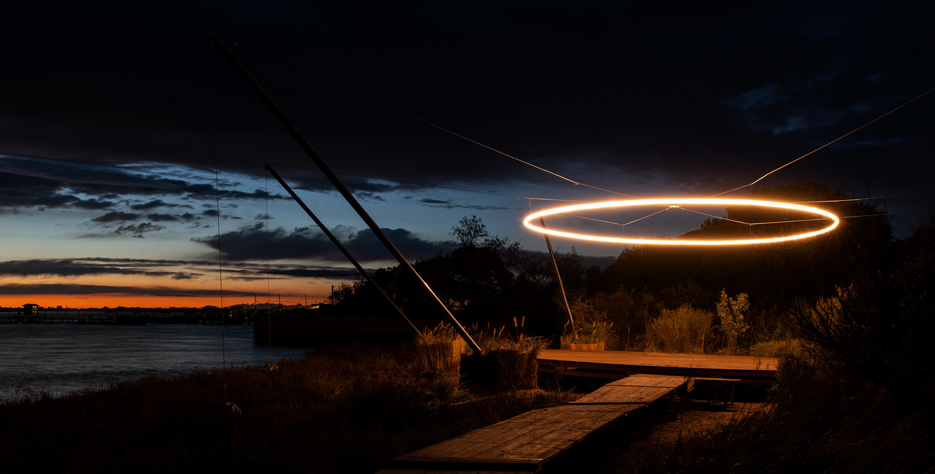 Glowing circular LED ring light installation suspended outdoors at dusk, reflected over waterfront with dramatic twilight sky