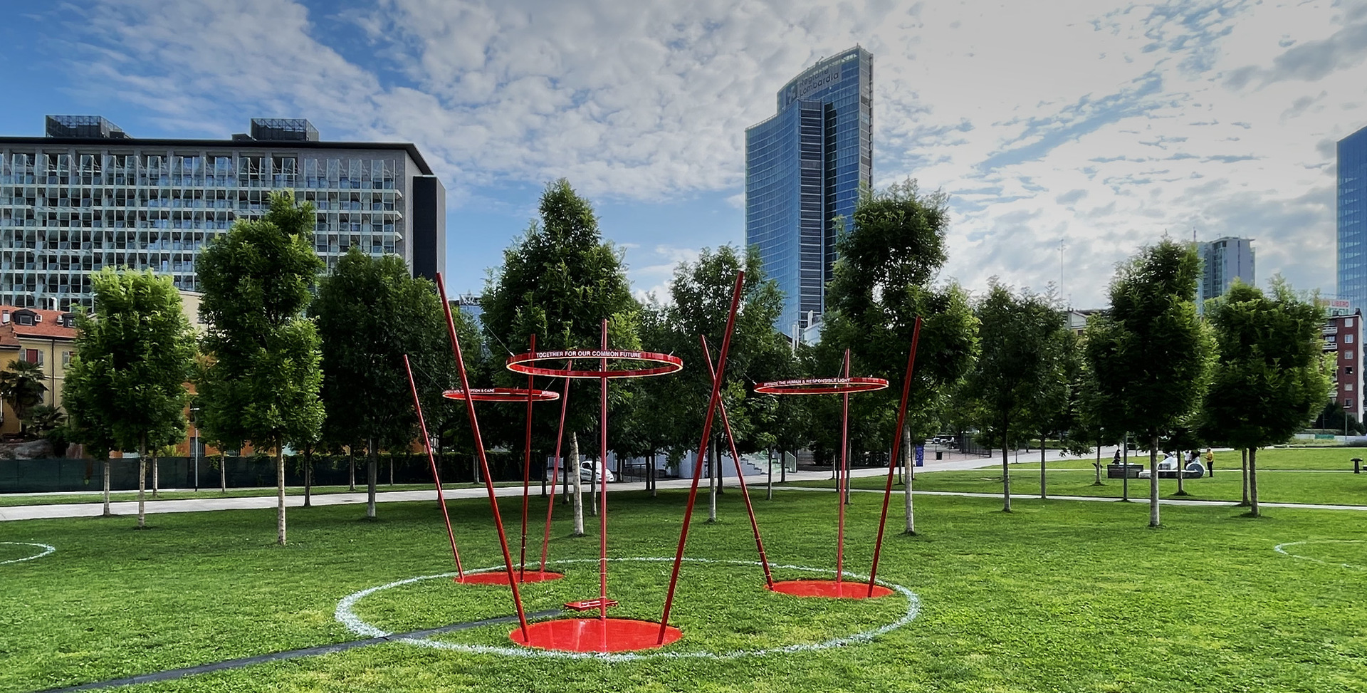 Red sculptural outdoor lighting installation with circular ring fixtures on angled poles, set on green lawn with urban skyline background.