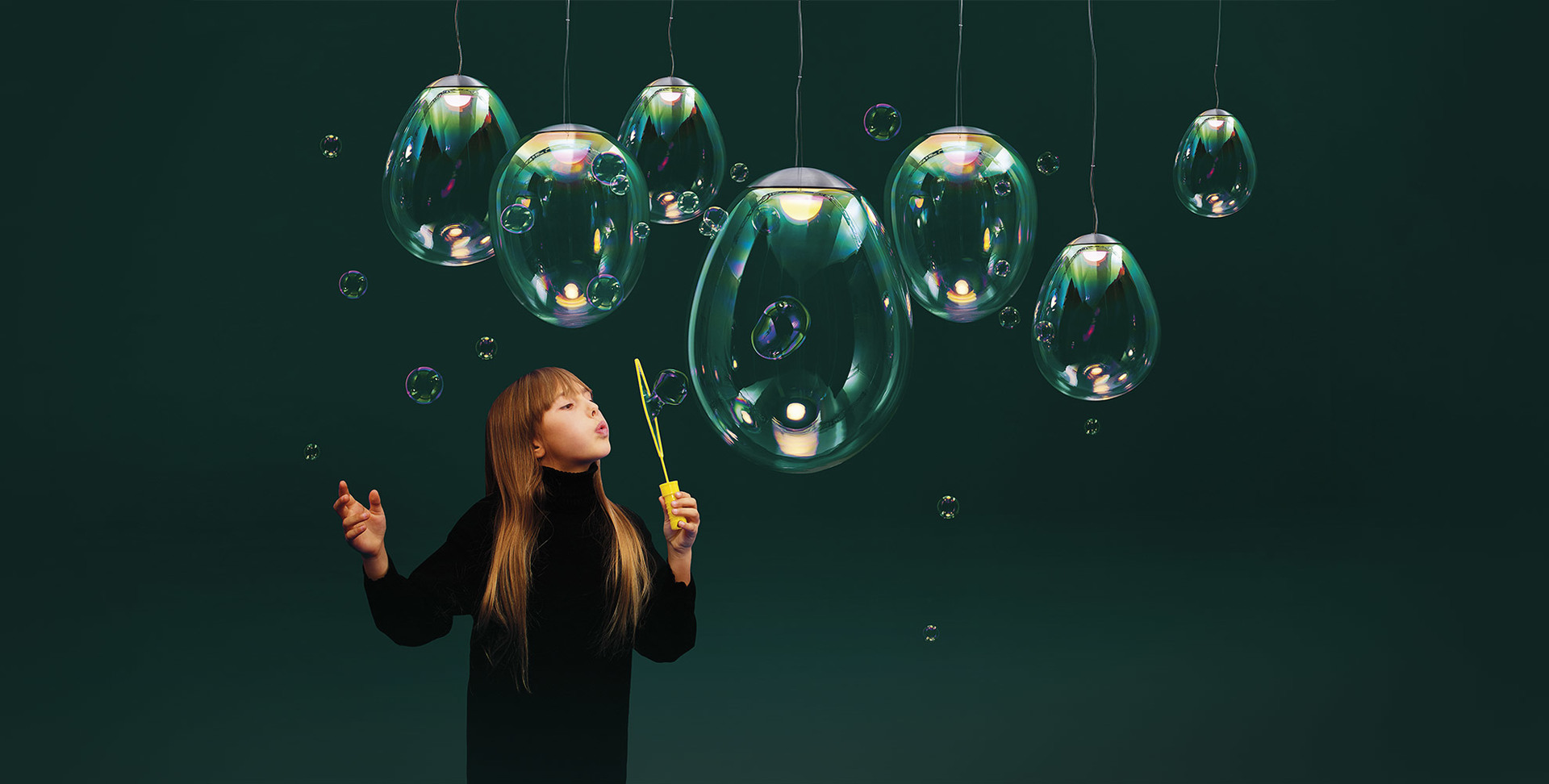Girl blowing soap bubbles near Artemide iridescent glass pendant lamps resembling floating bubbles against dark green background