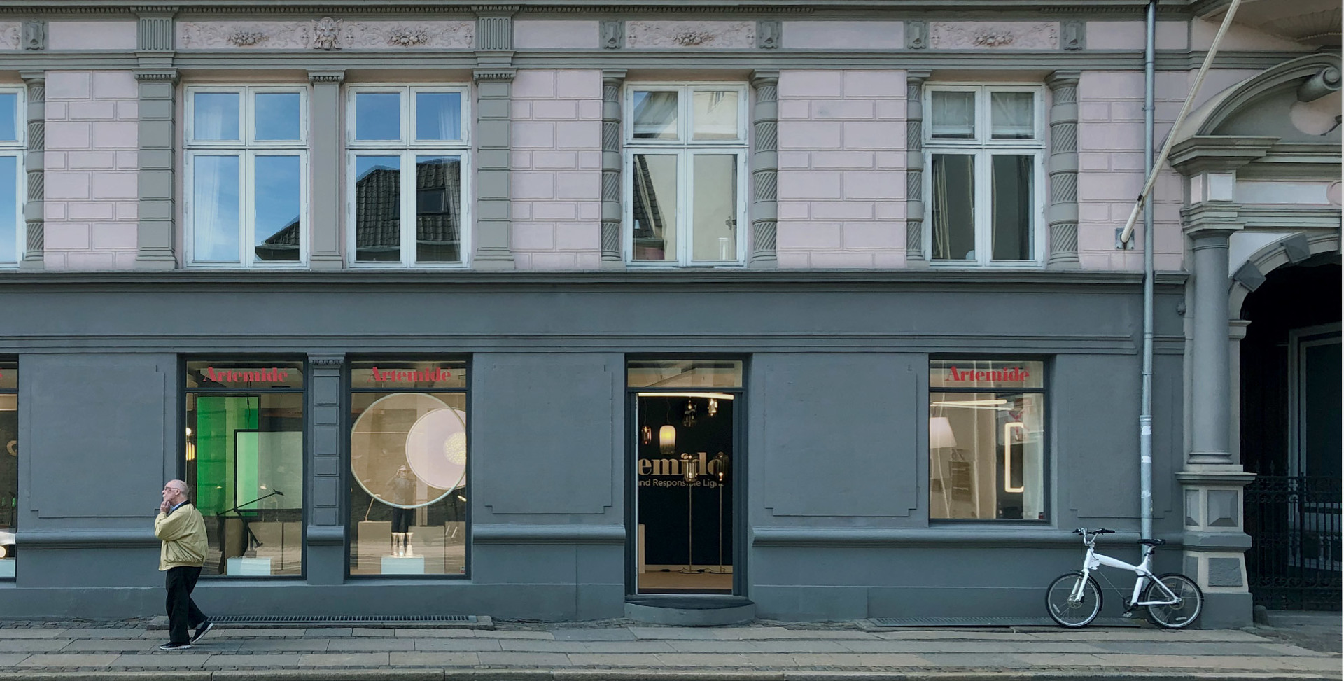 Artemide store exterior with dark grey facade, large display windows showcasing pendant lamps, street-level view with passerby and bicycle