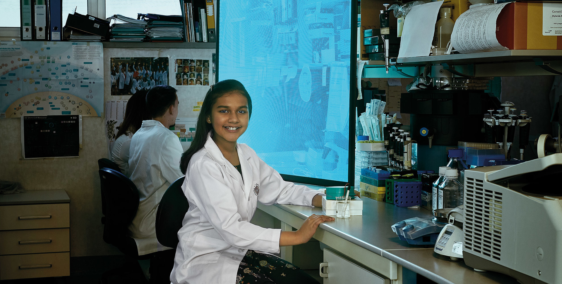 Young scientist in white lab coat smiling at camera in a laboratory, colleague working at computer in background