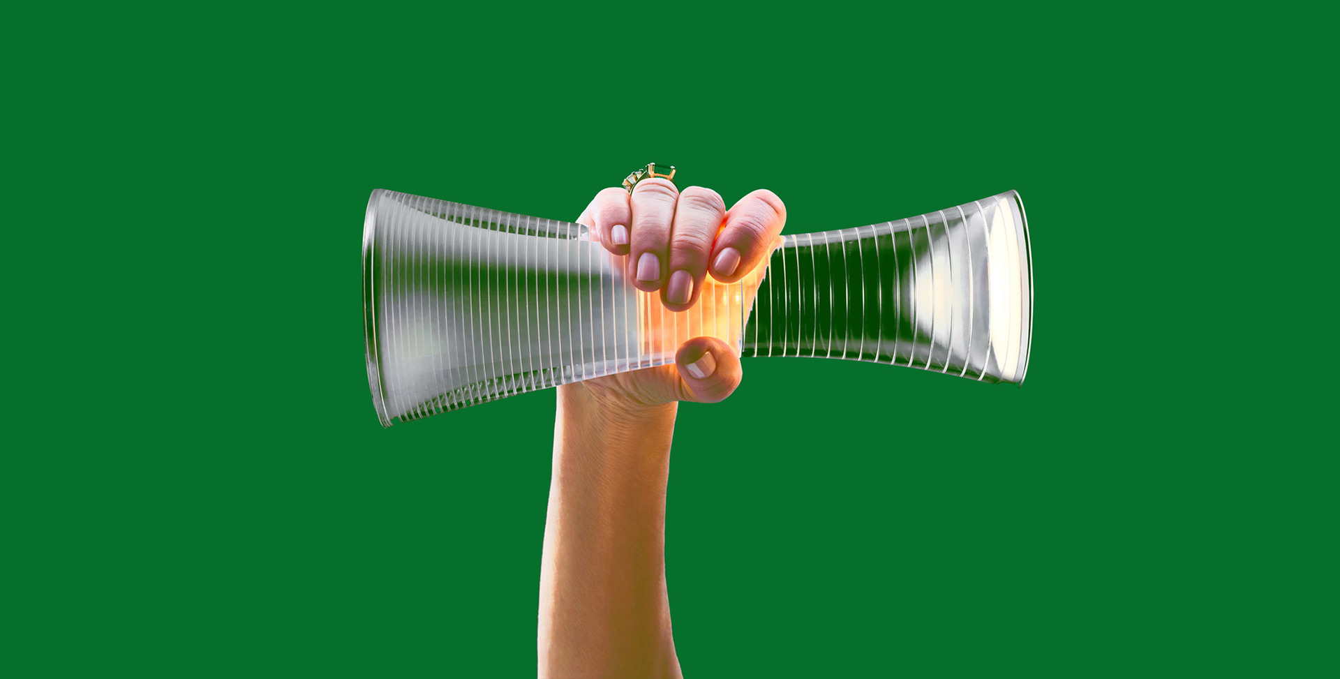 Hand holding an hourglass-shaped ribbed glass lamp diffuser glowing with warm light, against a green background