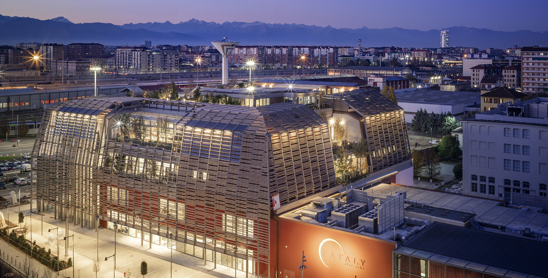 Aerial dusk view of Eataly Turin, a modern building with illuminated wooden lattice facade and warm interior lighting against Alpine skyline