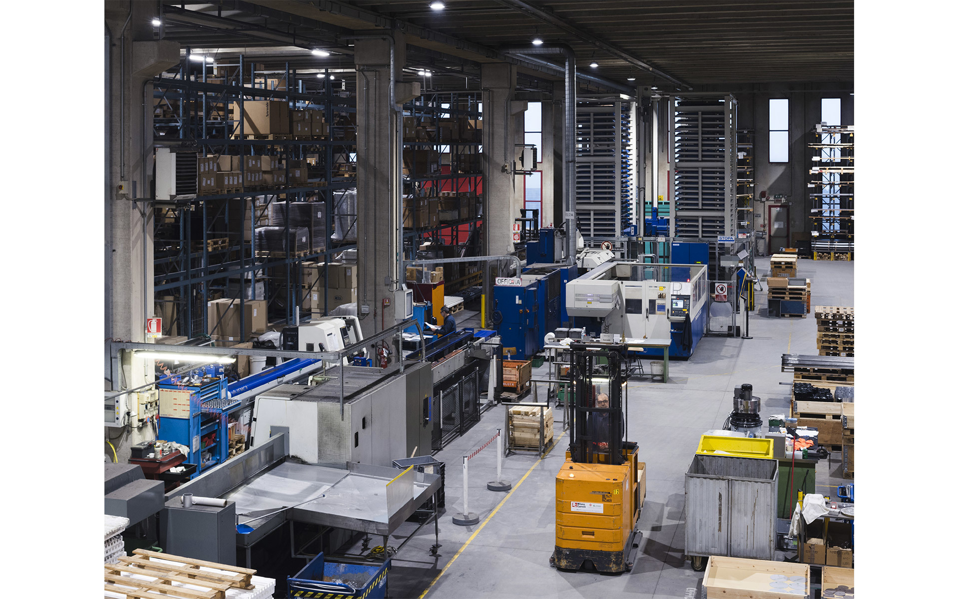 Large industrial facility with machinery, automated storage systems, a forklift, and overhead lighting illuminating the workspace.