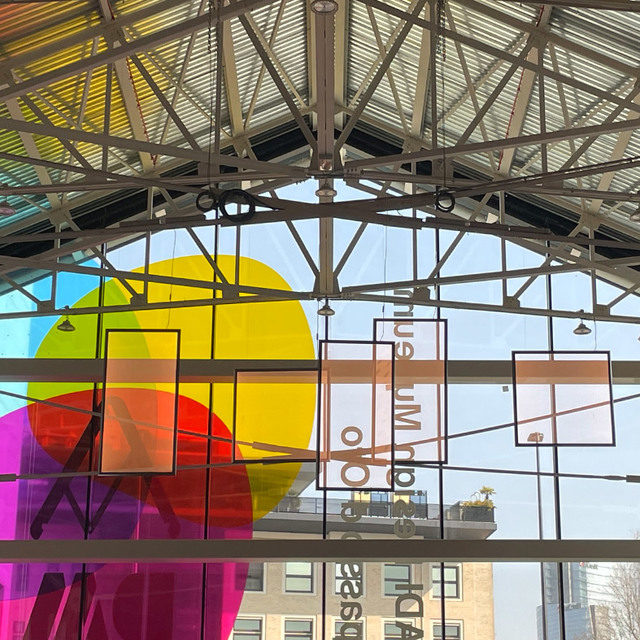 Glass-roofed atrium with steel trusses, colorful translucent geometric shapes and circles in yellow, red, and purple on large windows