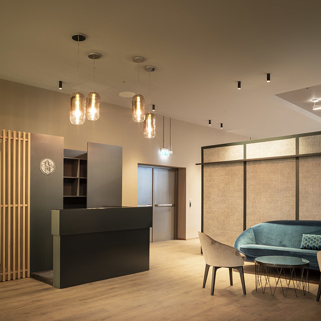 Modern reception area with amber glass pendant lights above a dark desk, slim ceiling spotlights, and a teal sofa in a lounge corner.