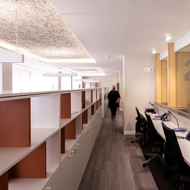 Modern office corridor with recessed ceiling lights and textured panels illuminating workstations and a reception desk along both sides.