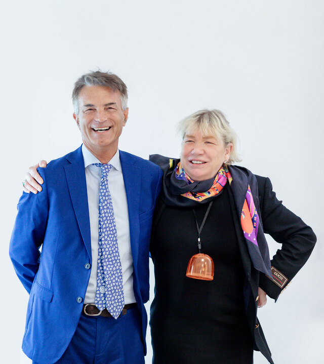 Two smiling adults posing together, a man in a blue suit and a woman in a black jacket with a colorful scarf