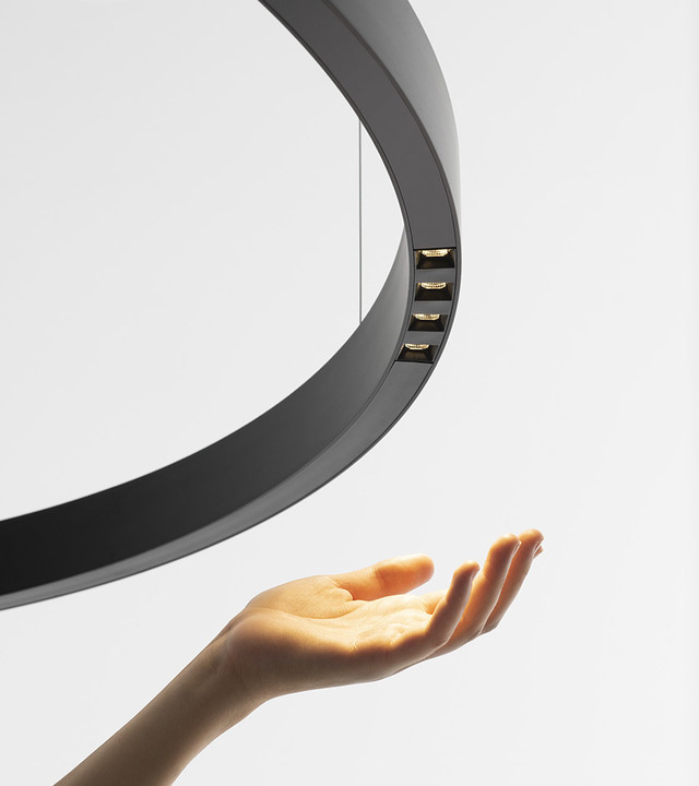 Circular black pendant lamp with downward LED light modules, with a hand reaching up beneath it.
