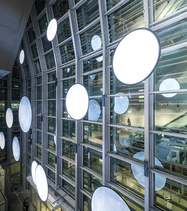 Multiple large circular LED pendant lights suspended in front of a multi-story glass curtain wall facade, creating a luminous installation.