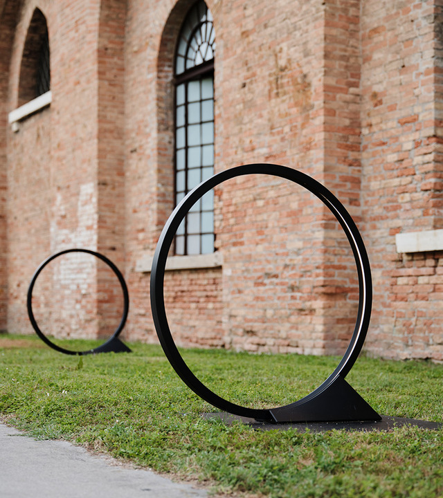 Two black circular ring outdoor floor lamps on grass, leaning against a historic brick building facade.