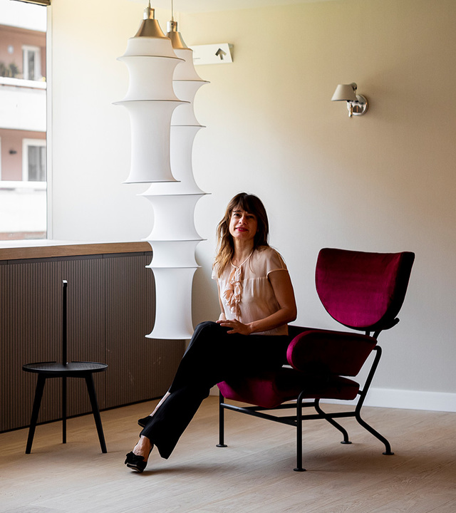 Woman seated in a red velvet armchair beside a tall white sculptural floor lamp and two brass pendant lights in a modern interior