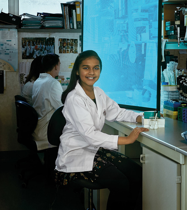 Young girl in a lab coat smiling at a laboratory workbench, with another student working in the background