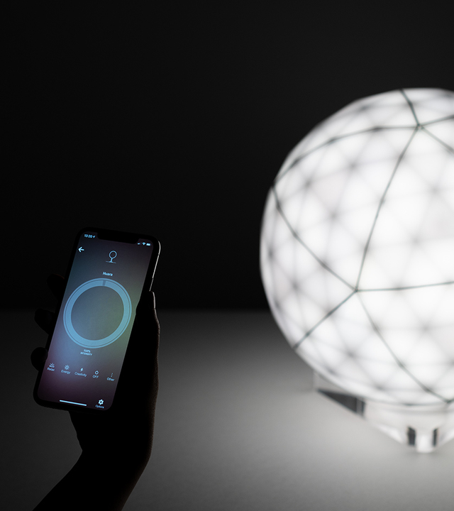 Hand holding a smartphone with Artemide app controlling a glowing spherical table lamp with geometric grid pattern diffuser.
