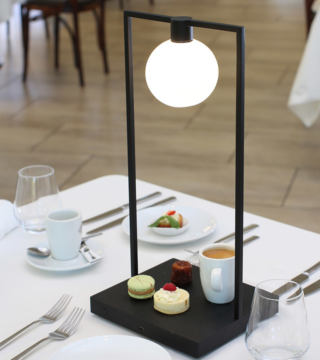 Artemide black rectangular table lamp with opal globe shade on dark base, displayed on a restaurant table with pastries and coffee.
