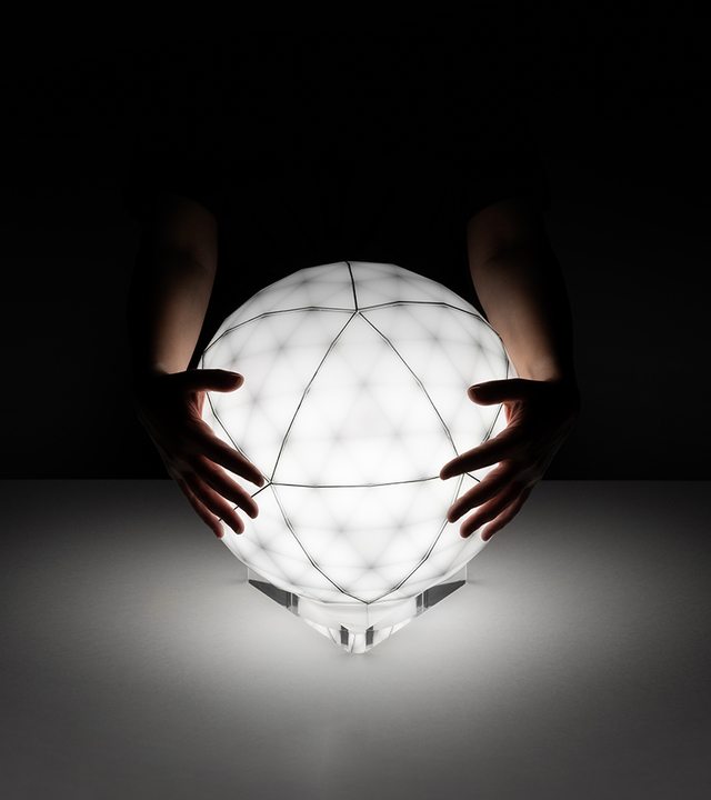 Hands cradling a glowing spherical lamp with geometric faceted surface on a transparent base, illuminating a dark surface.