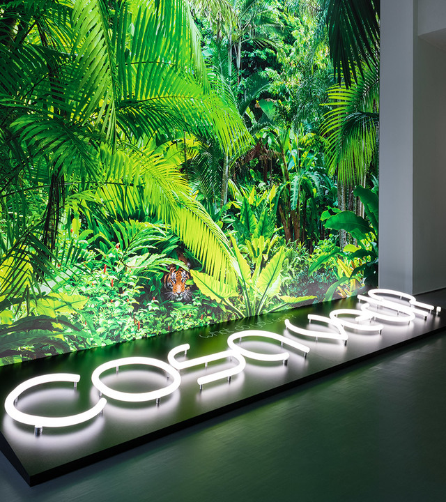 Glowing white neon letters spelling 'cosomeen' on a dark platform, set against a vivid tropical jungle mural with a hidden tiger.
