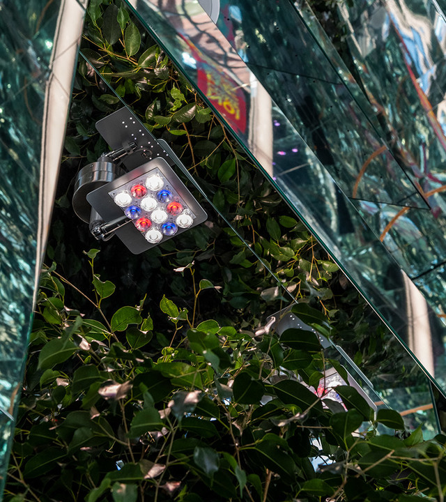Artemide outdoor LED spotlight with red, blue, and white LEDs mounted on a bracket among green foliage against a glass facade