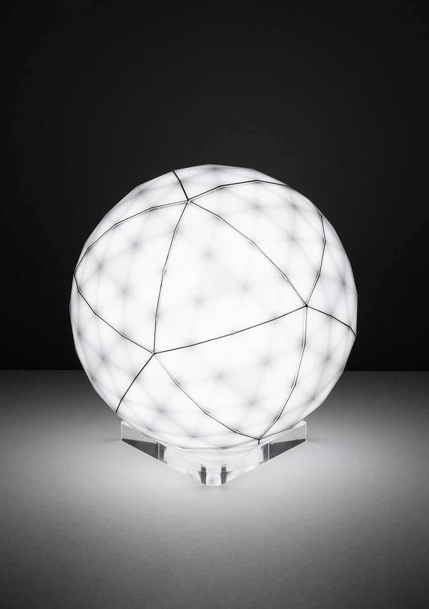 Artemide spherical table lamp with white diffuser, geometric black wire frame pattern, glowing evenly, on a transparent crystal base.