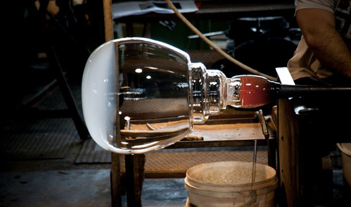 Glassblower shaping a large transparent glass globe for a lighting fixture on a metal blowpipe in a glassworks workshop
