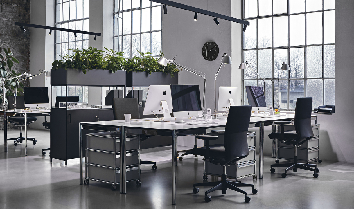 Modern open-plan office with Artemide Tolomeo desk lamps, workstations, black chairs, large windows, and green plant dividers