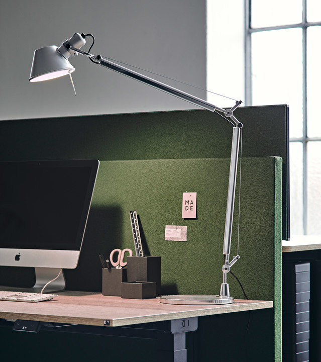 Artemide Tolomeo desk lamp in aluminum on a modern workstation, with adjustable arm extended over a monitor and green acoustic panel.
